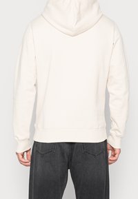 Sudadera con capucha color beige claro, con un bolsillo tipo canguro, puños y bajo acanalados, vista desde la parte posterior, combinada con jeans gris oscuro.