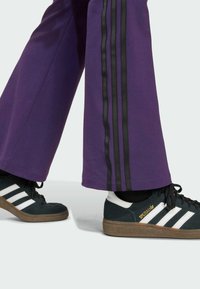 Purple sweatpants with black side stripes and loose fit, paired with black Adidas sneakers featuring white stripes and a brown sole.