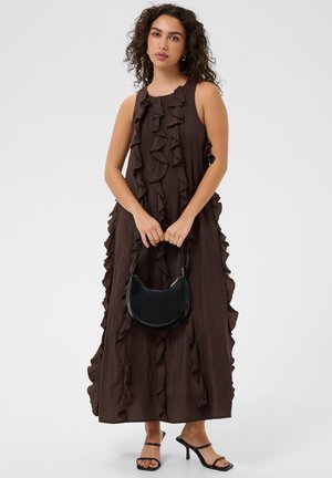 Femme aux cheveux bouclés portant une robe longue marron sans manches avec des volants, tenant un sac à main noir et portant des talons noirs à brides.