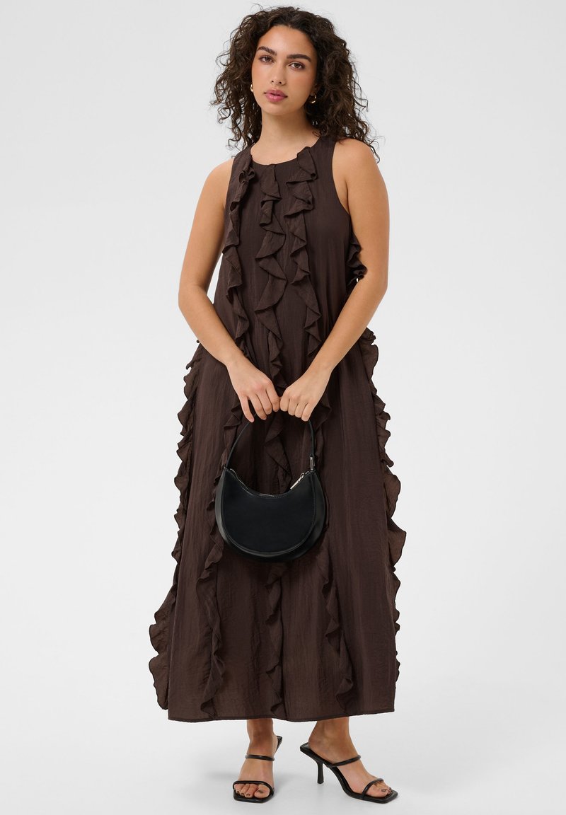 Femme aux cheveux bouclés portant une robe longue marron sans manches avec des volants, tenant un sac à main noir et portant des talons noirs à brides.
