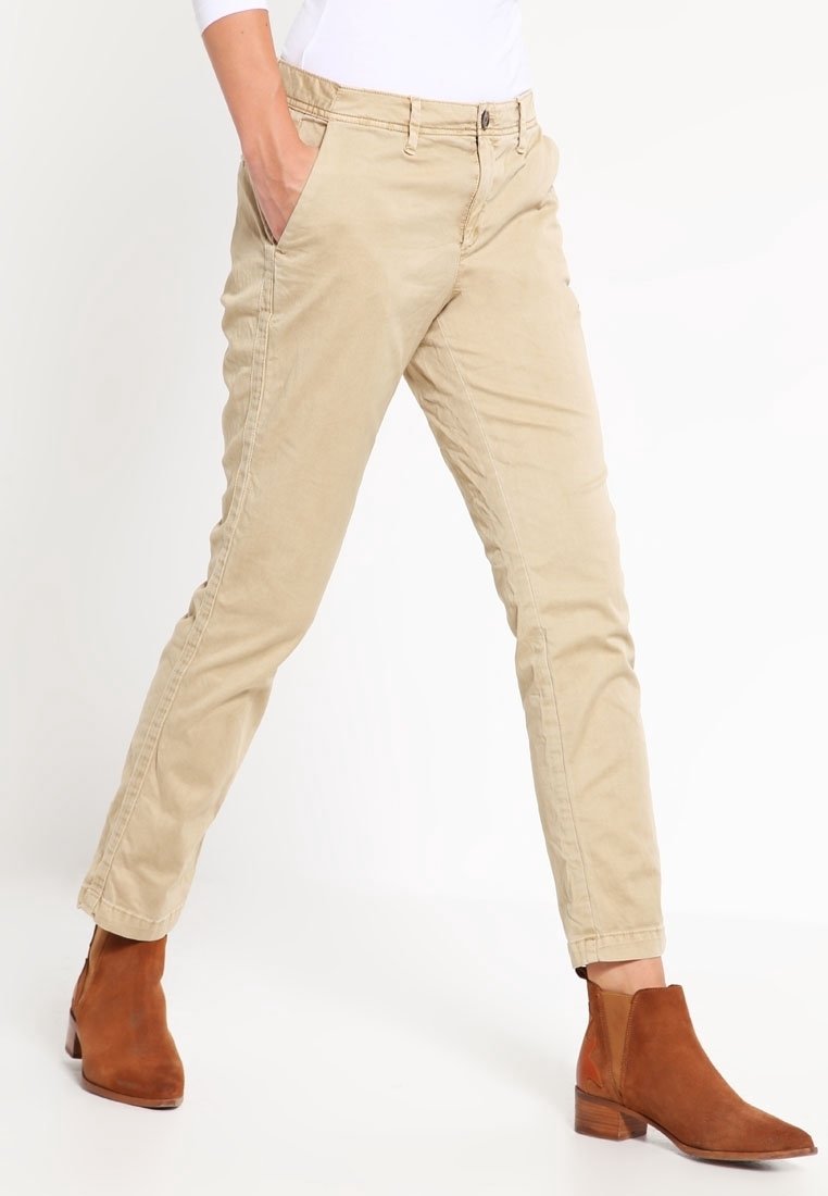 Chinos beige en coton avec une coupe slim, dotés de poches avant et d'une fermeture éclair, associés à des bottines brunes. Texture lisse, détails minimaux.