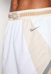 Hvite og beige sportsshorts med elastisk midjebånd, teksturert stoff, trekantet mønster og en grå Nike-logo på siden.