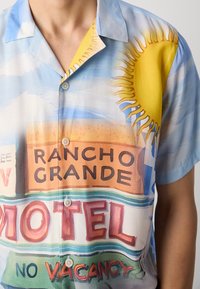 Majica s kratkimi rokavi, ki prikazuje barvito grafično sliko z soncem, oblaki in znamenji motelov. Narejena iz lahkega materiala z gladko teksturo.