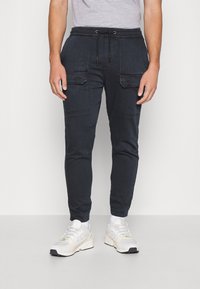 Laivastonsiniset collegehousut nyöritettävällä vyötäröllä, kahdella etutaskulla ja resoreilla nilkoissa, yhdistettynä valkoisiin urheilukenkiin.