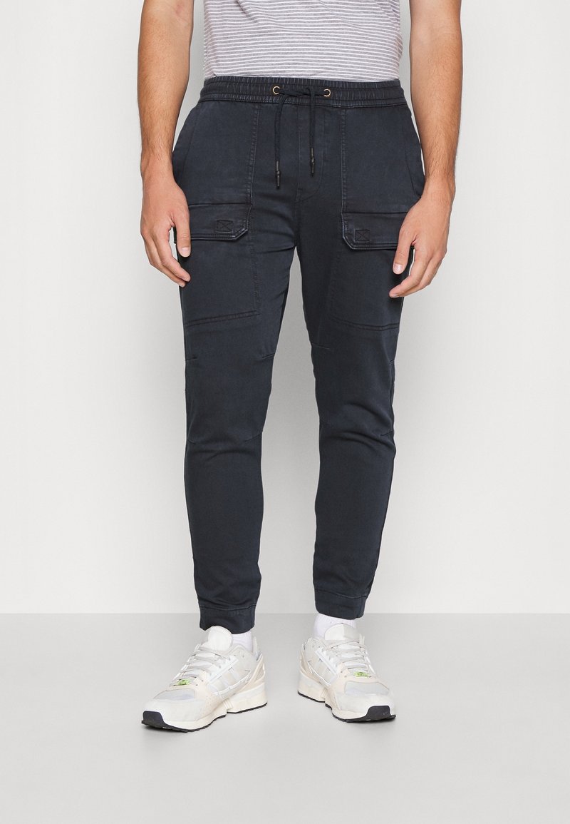 Laivastonsiniset collegehousut nyöritettävällä vyötäröllä, kahdella etutaskulla ja resoreilla nilkoissa, yhdistettynä valkoisiin urheilukenkiin.