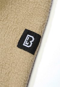Beige színű bolyhos anyag, puha textúrával, fekete címkével és fehér betűkkel, amely a szélehez van rögzítve.
