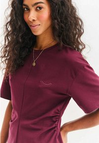 Bordowy t-shirt z miękkiej tkaniny z krótkimi rękawami, ozdobiony haftem rzęsy na lewej piersi oraz marszczeniem z boku.