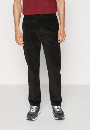 Pantaloni in corduroy neri con vestibilità dritta, caratterizzati da una texture sottile, tasche frontali e chiusura con bottone, abbinati a sneaker scure.