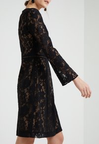 Kvinna klädd i en knälång svart spetsklänning med långa ärmar och blommönster, stående mot en enfärgad vit bakgrund.