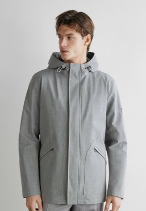 Junger Mann trägt eine hellgraue Kapuzenjacke mit Reißverschlusstaschen und blickt zur Seite vor einem schlichten hellen Hintergrund.