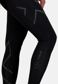 Czarne legginsy sportowe o obcisłym kroju, z refleksyjnymi szarymi akcentami i wzorzystymi elementami po bokach. Gładka tekstura.
