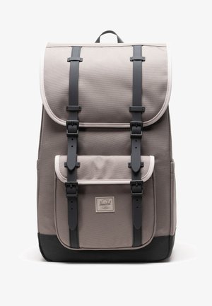 Zaino beige e nero con tasca anteriore, due cinghie nere verticali con fibbie e una maniglia superiore, su uno sfondo bianco.