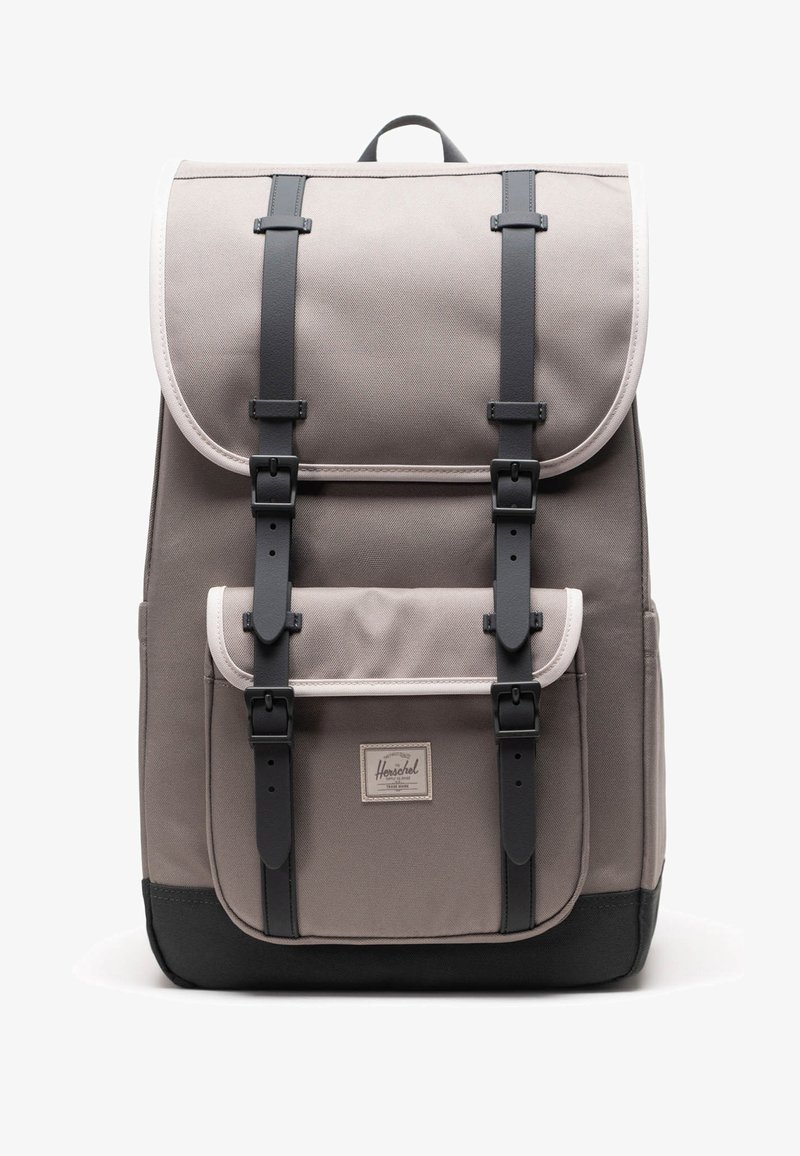 Bežovo-čierny batoh s predným vreckom, dvoma vertikálnymi čiernymi sponami a horným úchytom, na bielom pozadí.