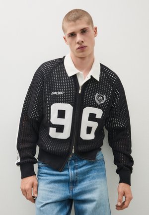 Junger Mann trägt eine schwarze Mesh-Zipjacke mit weißer Nummer 96 und hellblauen Jeans, stehend vor einem schlichten hellen Hintergrund.