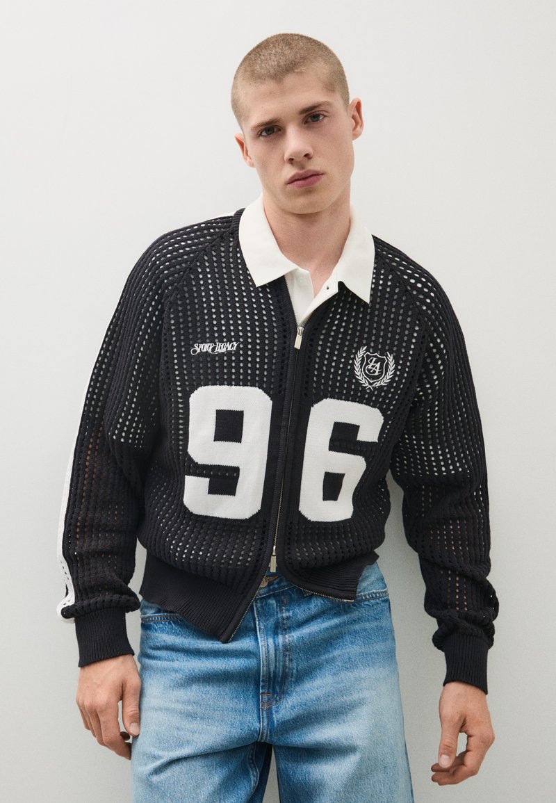 Jeune homme portant une veste zippée en maille noire avec le numéro blanc 96 et un jean bleu clair, debout devant un fond clair uni.