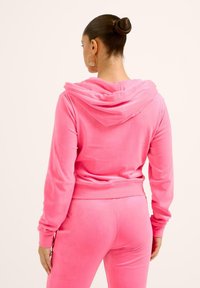 Rožinė velūrinė sportinė apranga su gobtuvu ir ilgomis rankovėmis, pasižyminti prigludusia fasonu. Audinys atrodo minkštas ir purumas prieš odą.