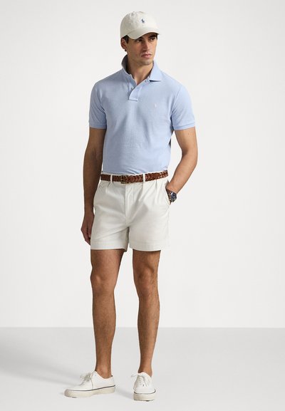 Polo de culoare albastru deschis, pantaloni scurți albi, curea împletită maro și adidași albi. Șapca are un mic logo. O ținută neată și lejeră pentru exterior.