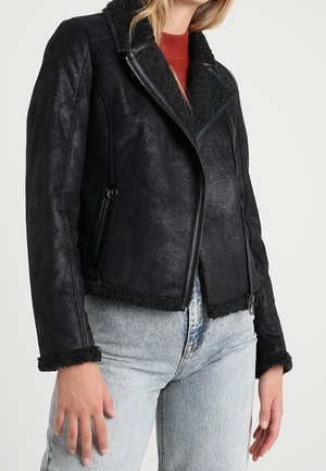 Kobieta w czarnej skórzanej kurtce z ociepleniem z baranka, z kieszeniami na zamek oraz jasnoniebieskich dżinsach, stojąca na jednolitym tle.