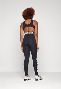 Sutien sport negru cu spate tăiat, asortat cu colanți negri cu talie înaltă, având text alb "NEBBIA" pe picior. Pantofii sport completează ținuta.