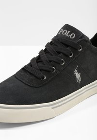 Svarta mockaskor med svarta snören, broderad logotyp på sidan, gräddfärgad gummisula med subtila ränder, och metalliska öglor.