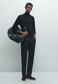 Schwarzer Rollkragenpullover, maßgeschneiderte schwarze Hose und dunkelbraune Schuhe. Trägt eine große, weiche schwarze Leder-Reisetasche mit kurzen Griffen.