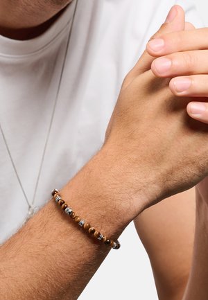 Mann trägt weißes Hemd und Perlenarmband mit braunen und silbernen Perlen, hält die Hände nahe an der Brust zusammen.