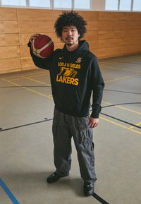 Moletom preto com texto amarelo "Los Angeles Lakers", calças cargo cinzentas e ténis pretos. A segurar uma bola de basquete, numa ginásio com paredes de madeira.