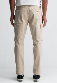 Man in beige cargo broek en zwarte sneakers staand tegen een plain witte achtergrond, gezien van achteren.