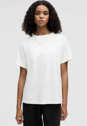T-shirt blanc à manches courtes en coton, avec une coupe décontractée et un col rond, porté avec un pantalon noir. Texture simple et lisse.