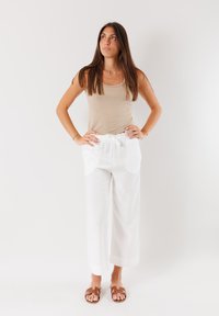 Top a maniche lunghe beige abbinato a pantaloni larghi bianchi, leggeri, con vita elastica e tasche. Sandali marroni con cinturini ai piedi.