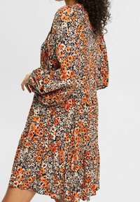 Blumenkleid mit einem orange-schwarzen Muster, langen Ärmeln, einem rüschengesäumten Saum und einem leichten Stoff für eine fließende Silhouette.