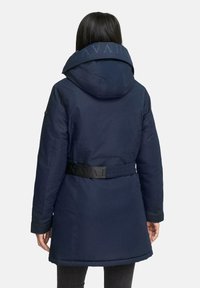 Parka longue bleu marine avec capuche, dotée d'une taille ceinturée et d'un tissu texturé. Des lettres sur le col ajoutent un détail de design.