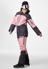 Tuta da sci nera e rosa chiaro con cappuccio, numerose tasche con zip e design aderente. Presenta un tessuto esterno testurizzato e dettagli neri.