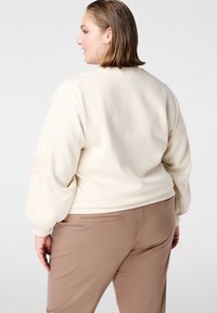 Crèmekleurige trui met gestructureerde pofmouwen en bloemenborduursel op de linkermouw, gecombineerd met taupekleurige broek, gezien van achteren.