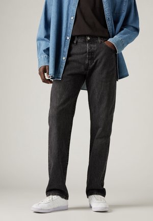 Person trägt schwarze Jeans, weiße Sneakers, schwarzes Hemd und offene blaue Jeansjacke mit einer Hand in der Tasche.