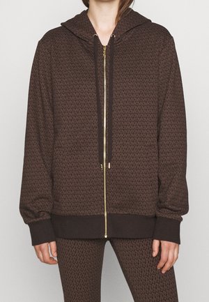Personne portant une veste à capuche marron zippée et un pantalon assorti avec un motif MK répétitif.