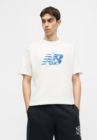 Mladý muž v bielom tričku s modrým logom „NB“ a čiernych šortkách s textom „New Balance“, ruky v vreckách, pozerá sa do strany.