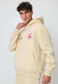 Jeune homme aux cheveux bouclés portant un sweat à capuche crème avec un motif de fleur rose et un texte sur la poitrine, posant devant un fond uni.