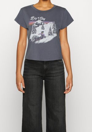 Woman wearing a dark cropped t-shirt with a skier and pine trees graphic and "Big Sky" text, paired with high-waisted black jeans.