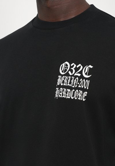 T-shirt noir avec du texte gothique blanc sur la poitrine disant "032C Berlin-2001 Hardcore" porté par une personne.