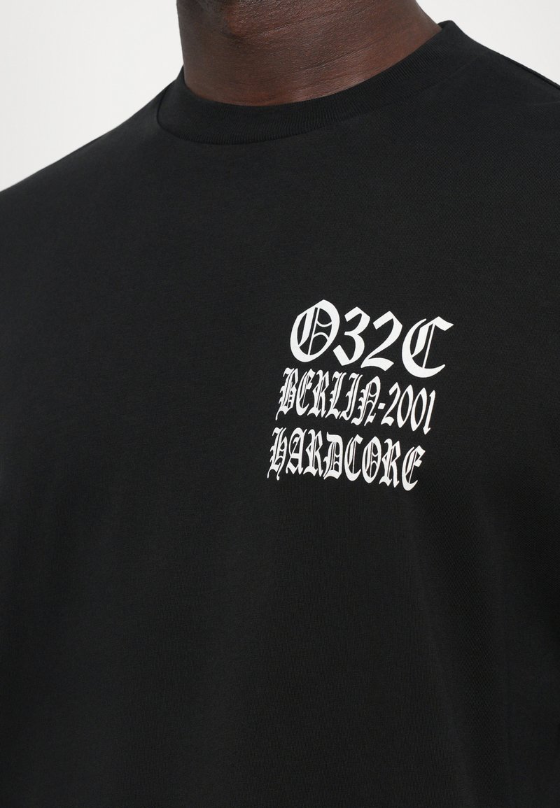 Black T-shirt with white Gothic text on chest reading "032C Berlin-2001 Hardcore" worn by person.