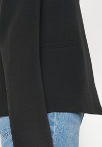 Černý blazer s hladkou texturou, detail jednolité kapsy a otevřeným předním dílem, kombinovaný se světle modrými džíny.