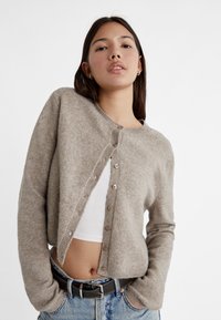 Light brown, knitted cardigan with a round neck, button closure, and ribbed cuffs. Worn over a white top, paired with blue jeans.