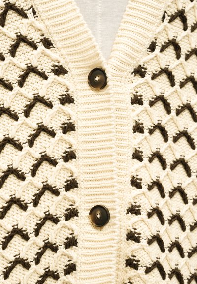Κρεμ πλεκτή ζακέτα με μαύρο ζιγκ-ζαγκ μοτίβο και δύο μεγάλα σκουρόχρωμα κουμπιά στο εμπρόσθιο μέρος.