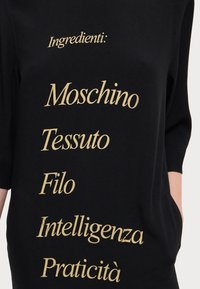 Fekete szövet felső, arany színű felirattal, amely az alábbiakat sorolja fel: „Hozzávalók: Moschino, Tessuto, Filo, Intelligenza, Praticità.” Rövid ujjak, laza fazon.