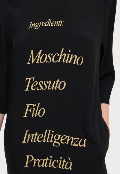 Black fabric top featuring gold text listing “Ingredients: Moschino, Tessuto, Filo, Intelligenza, Praticità.” Short sleeves, relaxed fit.