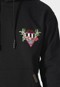 Felpa nera con cappuccio caratterizzata da un design grafico colorato che presenta foglie di palma, una macchina da corsa e un motivo a scacchi nelle tonalità di rosa, rosso e verde.