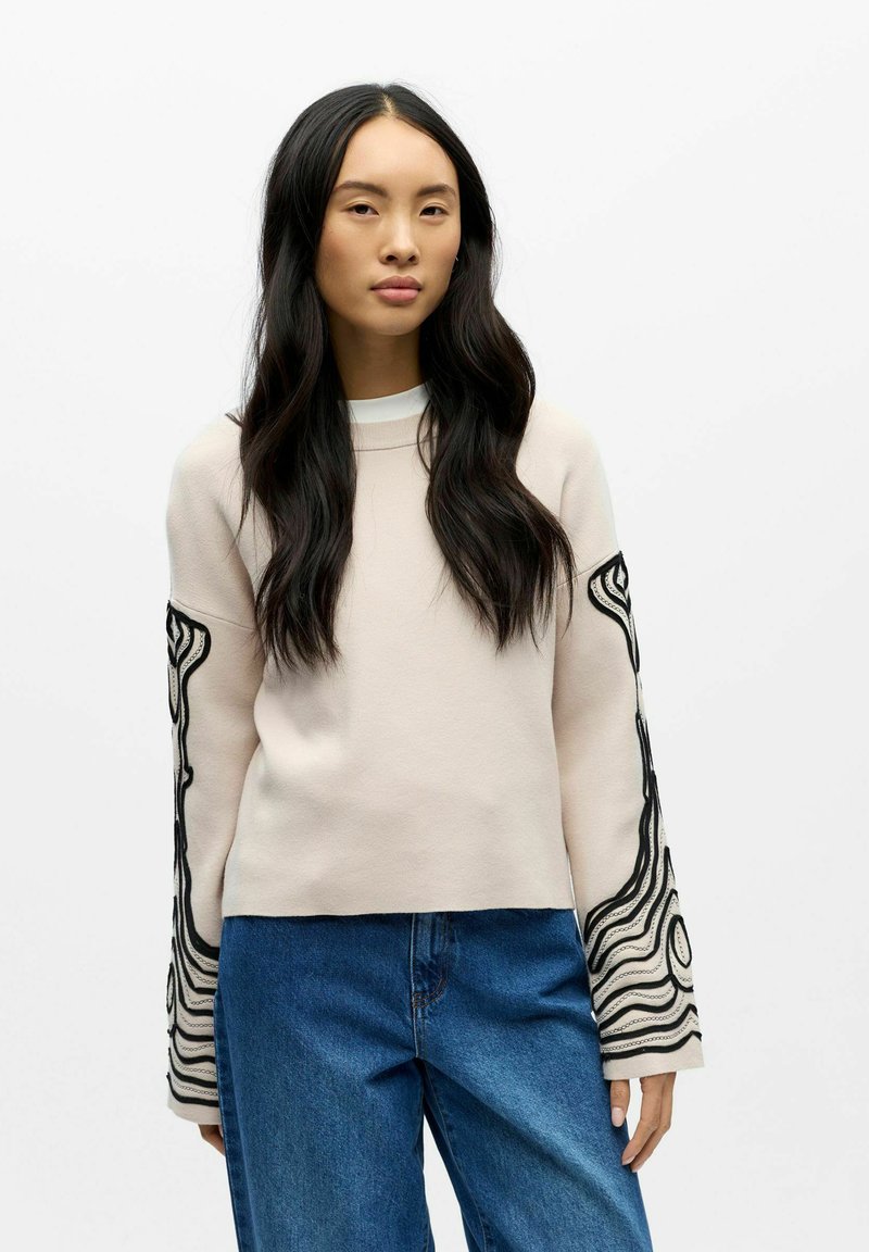 Hellrosa Sweatshirt mit langen Ärmeln und schwarzer abstrakter Stickerei. Weicher Stoff, Rundhalsausschnitt, lockere Passform, kombiniert mit blauer Jeans.