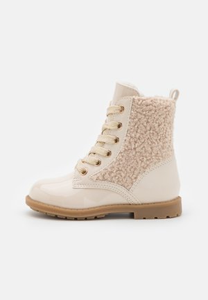 Botas de tornozelo bege com um bico em couro laqueado brilhante e atacadores, tecido texturado e felpudo na parte superior do cano, e sola em borracha.
