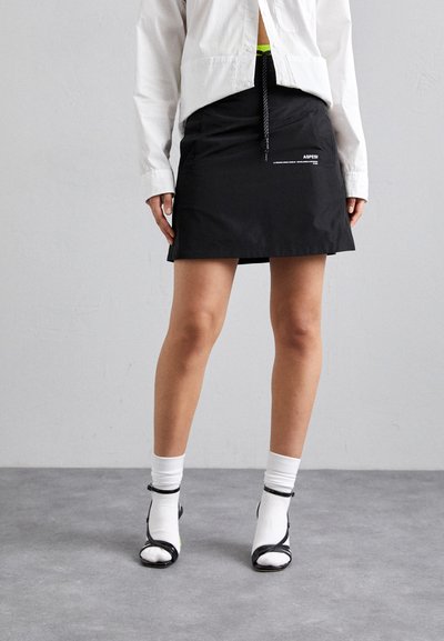 Jupe noire avec une taille à cordon, accentuée par un petit logo blanc. Assortie à des chaussures noires à bout ouvert et des chaussettes blanches mi-mollet. Texture lisse.